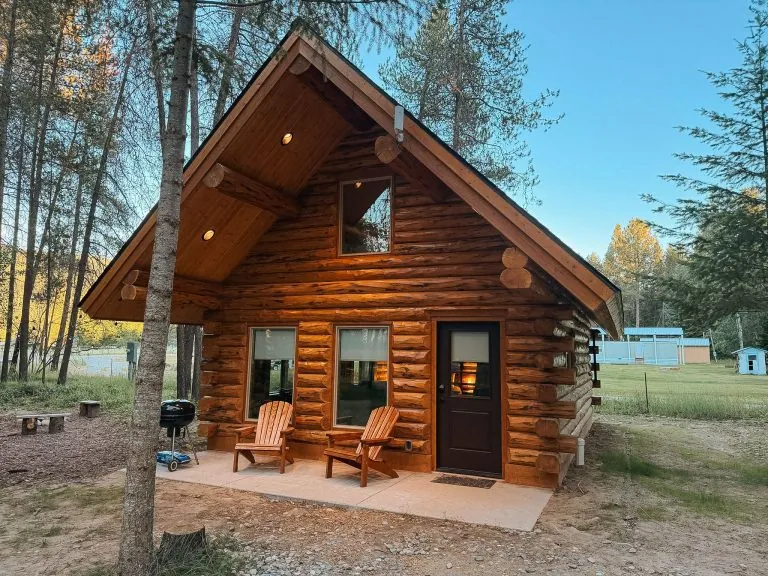 Front view of compact log cabin with wood patio, Adirondack chairs, and tall pine trees